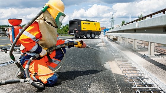 Oberflächenvorbereitung an einer Autobahnbaustelle – mobiler KAESER Kompressor versorgt Druckluftwerkzeuge zur Reinigung und Behandlung von Metallteilen.